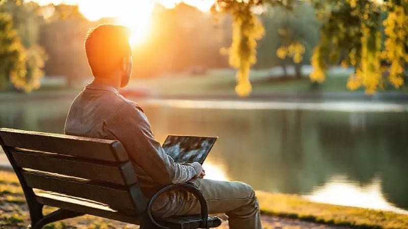 A photorealistic image of a person sitting peacefully on a park bench, looking at a serene lake, with a laptop closed beside them. The setting sun casts a warm, cinematic light, emphasizing tranquility and disconnection from work. Sharp focus on the person, depth of field blurring the background, 8K hyper-detailed, professional photography, shot on a high-end DSLR.